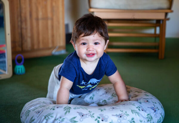 A baby boy in a dinosaur t-shirt grins at the camera while propped up on a nursing pillow on a green carpeted floor.