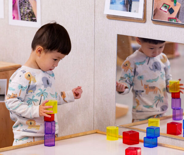 A toddler in a patterned sweatshirt concentrates on stacking colorful translucent blocks at a table, with his reflection visible in a mirror behind the blocks. Photos of children playing are displayed on the wall above.