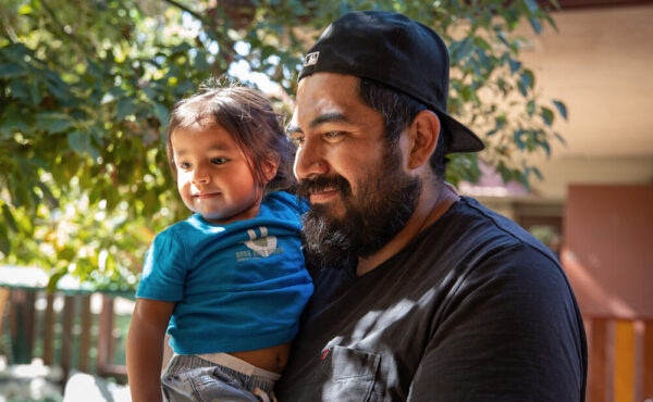 A bearded father holds his young toddler in his arms outdoors, both looking off to the side in dappled sunlight beneath tree branches.